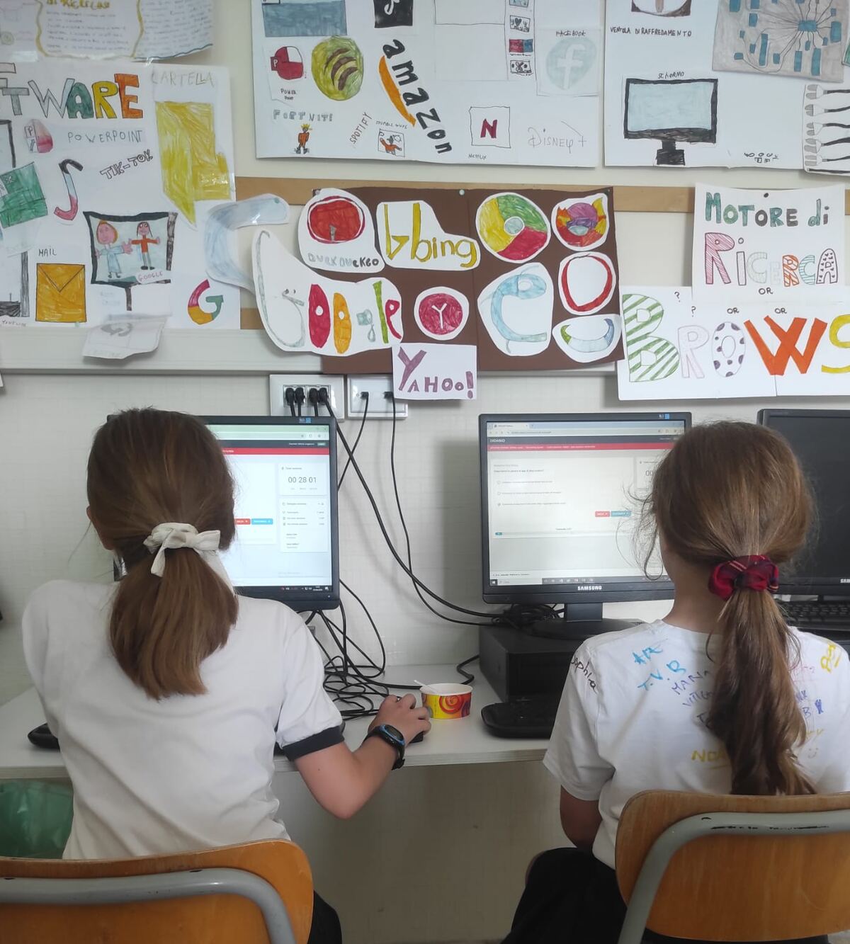 Due alunne al computer nel laboratorio di informatica durante una prova EIPASS, con i cartelloni della classe sul digitale alle pareti